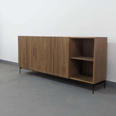 Vista a 45° hacia la izquierda del mueble vajillero color madera Peteribi, con cuatro puertas con tapa varilladas, y un espacio hueco con estante al medio en su parte derecha. Tiene cuatro patas metálicas.