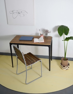 Vista de frente superior del mueble escritorio, que tiene tapa de madera y estructura de hierro, cuatro patas al piso. Ambientado con una silla y libros.