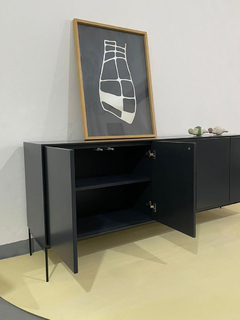 Vista de frente con inclinación hacia la derecha del mueble vajillero, color gris oscuro, que tiene cinco puertas (dos abiertas, las primeras de la izquierda donde se ve un estante) y cuatro patas metálicas, ambientado en un living.