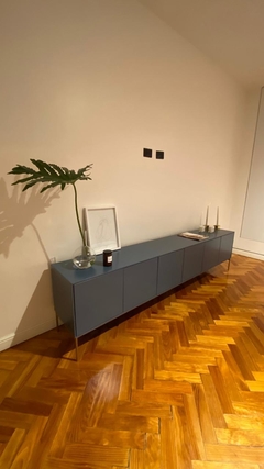Vista a 45° hacia la derecha del mueble consola, color azul, que tiene seis puertas y cinco patas metálicas al piso, ambientado en un living.