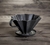 1 coador de café em cerâmica preto, está apoiado sobre uma mesa de madeira 