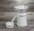 Vista de perfil de um Kit de Jarra e Coador de Café V60-2 em Cerâmica com esmaltação branca da Felline Cerâmica. O Coador está encaixado na jarra, ao lado tem um pequeno corta pingos também na cor branca. O conjunto está sobre uma mesa rústica de madeira.