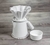 Kit de Jarra e Coador de Café V60-02 em Cerâmica com esmaltação branca da Felline Cerâmica. O Coador está encaixado na jarra, ao lado tem um pequeno corta pingos também na cor branca. O conjunto está quase de perfil sobre uma mesa rústica de madeira.