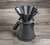 Vista de perfil de um Kit de Jarra e Coador de Café V60-02 em Cerâmica com esmaltação preta da Felline Cerâmica. O Coador está encaixado na jarra, ao lado tem um pequeno corta pingos também na cor preta. O conjunto está sobre uma mesa rústica de madeira.