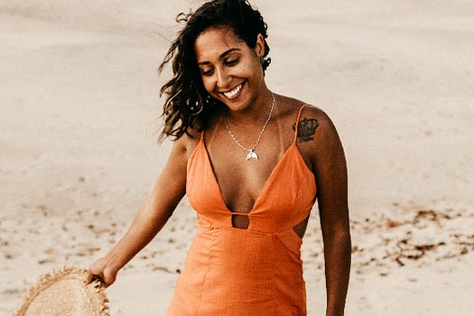 mulher morena bronzeada caminhando pela praia usando um vestido de alça decotado na cor
    laranja