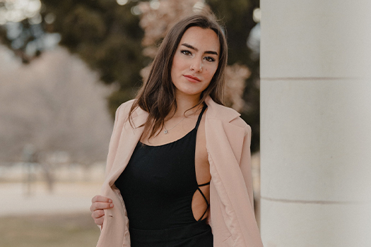 Mulher posa com blusa regata preta com fenda lateral e blazer bege por cima aberto.