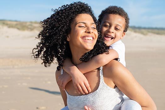 Mulher negra com cabelos cacheados ao vento carrega filho nas costas enquanto sorri.