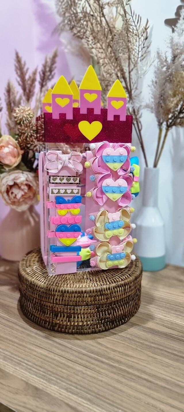 Porta laços em acrílico transparente com decoração de castelo nos tons de rosa escuro, rosa claro e amarelo, imagem com laços pequenos e hair clips presos na fita do porta laços