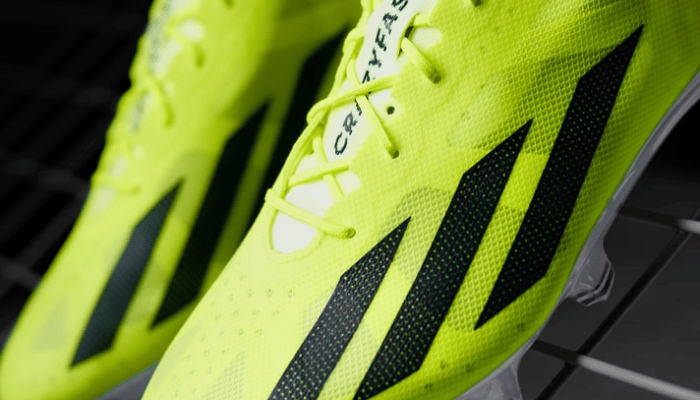 Chuteira de Campo Adidas X Crazyfast+ FG Solar Energy. A chuteira é predominantemente verde com detalhes em laranja neon. Possui um cabedal sintético leve e texturizado para melhor controle de bola.  Três listras pretas da Adidas estão estampadas na lateral. A chuteira tem uma sola com vários cravos para aderência em campos de grama natural.