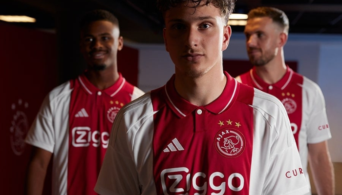 camisa titular do Ajax para a temporada 2024-2025, produzida pela Adidas. O manto apresenta um design clássico, com a cor branca predominante e uma faixa vertical vermelha. A gola polo é vermelha com um botão branco. O logo da Adidas e o escudo do Ajax estão presentes no peito, ambos em branco. 