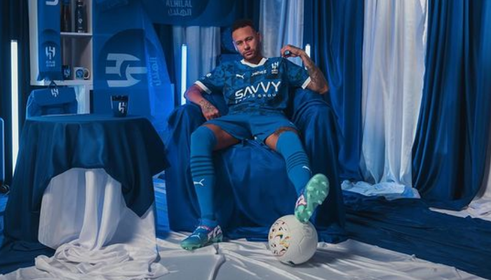 camisa titular do Al-Hilal para a temporada 2024-2025. A camisa é predominantemente azul, com um padrão de triângulos em tons da mesma cor na parte frontal e nas mangas. A gola é V, na cor branca, e o logo da PUMA aparece em branco nas mangas e no lado direito do peito.