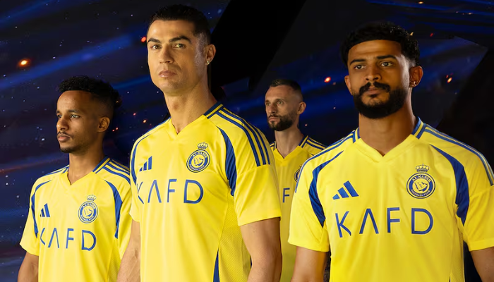 A camisa é predominantemente amarela, com detalhes em azul, como as três listras nos ombros, os recortes nas laterais e a gola V. O escudo do Al-Nassr é aplicado no lado esquerdo do peito, e a frase "Nassr runs in my blood" está presente na parte inferior esquerda da camisa.