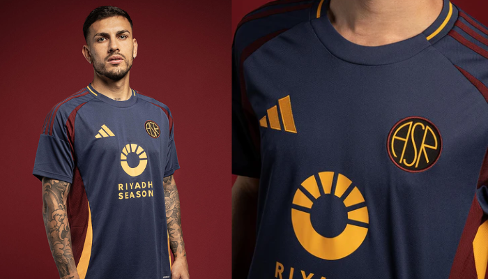 nova terceira camisa da AS Roma da Adidas. A camisa é predominantemente azul marinho e apresenta um escudo retrô circular com as siglas "ASR" no centro. Detalhes em bordô e amarelo, como as três listras nos ombros, recortes nas laterais e a numeração nas costas com textura inspirada no pavimento "sampietrini", completam o design.