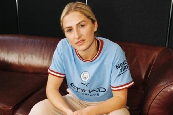 Camisa Feminina Manchester City Puma 2022/23 em azul celeste, com detalhes bordô, gola redonda e punhos únicos. Homenagem a Collin Bell, selo especial e logo Puma. Tradição e estilo retro exclusivo.