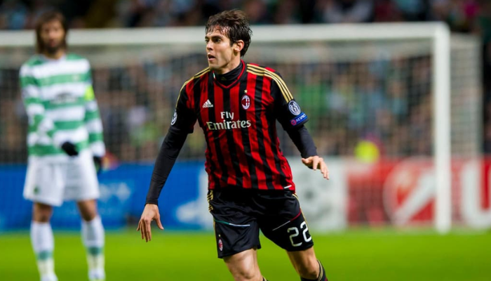 Camisa titular do AC Milan da temporada 2013/2014, exibindo o design clássico em vermelho, preto e dourado. A camisa apresenta o logo da Adidas e o patch da Serie A. É possível ver o nome "kaka" estampado nas costas.