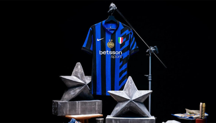 nova camisa titular do Inter de Milão para a temporada 2024/25. O manto, nas cores azul e preto, apresenta um design moderno com faixas diagonais que se cruzam com as tradicionais listras verticais. O escudo do clube, em dourado, destaca-se no centro da camisa, acompanhado da segunda estrela.