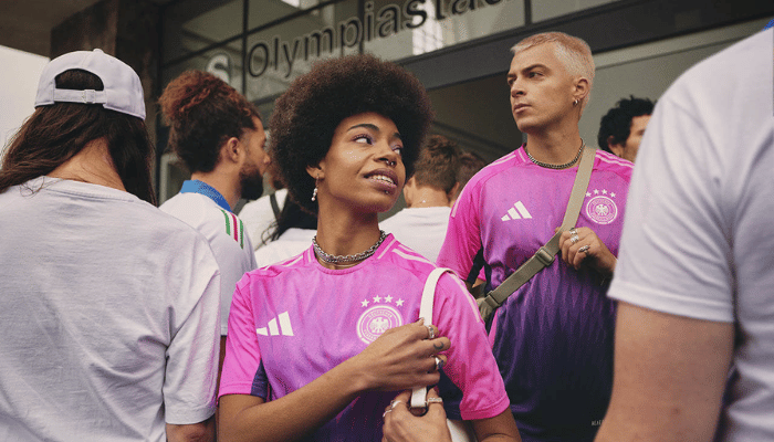 Camisa da seleção alemã com degradê de magenta para roxo. Gola redonda magenta com friso rosa. Três listras da Adidas nos ombros na cor rosa. Recortes curvos nas laterais. Escudo da DFB em magenta com detalhes brancos no lado esquerdo do peito.