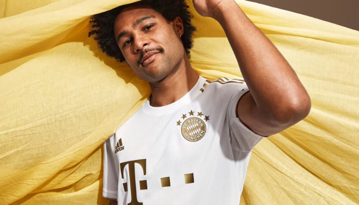 A Camisa Reserva Bayern de Munique Adidas 2022-2023 apresenta um design sofisticado com detalhes dourados, incluindo as listras nos ombros, punhos dourados e o escudo do clube monocromático em dourado.