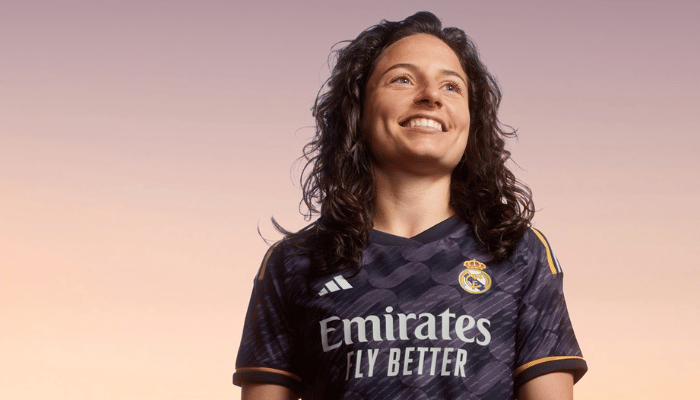 Camisa Reserva Feminina Real Madrid 2023-2024 Adidas. Camisa azul marinho com estampa de ondas, gola redonda, escudo do Real Madrid no lado esquerdo do peito, logo da Adidas no lado direito e três listras nos ombros nas cores amarelo, branco e roxo. 