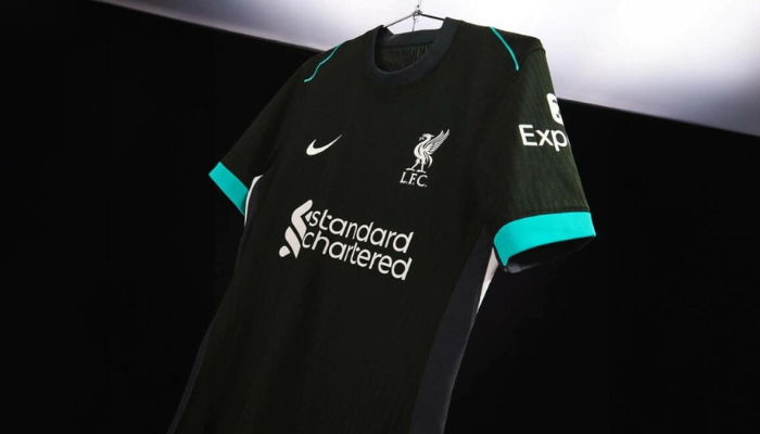 camisa reserva do Liverpool FC para a temporada 2024-2025. A peça, predominantemente verde escura, possui detalhes em verde "revisado" nos punhos e finas linhas nos ombros. As laterais do corpo apresentam listras em cinza com detalhes brancos. 