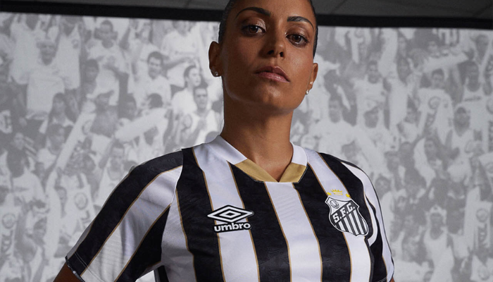 camisa reserva do Santos Futebol Clube 2024-2025, fabricada pela Umbro. A camisa apresenta listras verticais em branco e preto com bordas douradas, detalhes gráficos que homenageiam a Vila Belmiro, gola V com parte frontal dourada e nuca branca, e punhos listrados com referência ao estádio.