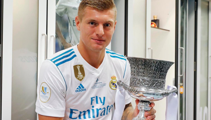 Camisa titular retro do Real Madrid Adidas 2017-2018 na cor branca e azul. A camisa possui o escudo do Real Madrid no lado esquerdo do peito, as três listras da Adidas nos ombros e o logotipo da Champions League na manga direita. Nas costas, o nome "Real Madrid" está impresso em branco acima do número 12. A camisa é usada por jogadores como Cristiano Ronaldo, Gareth Bale e Sergio Ramos.