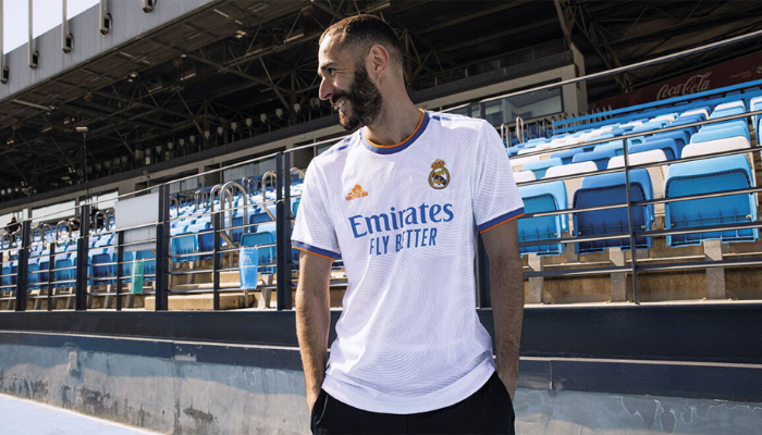 Camisa titular retro do Real Madrid para a temporada 2021-2022. A camisa branca apresenta gola redonda e punhos canelados em azul e laranja, com finas listras laranjas. As três listras da Adidas nos ombros são azuis, enquanto o logo da Adidas no lado direito do peito é laranja. 