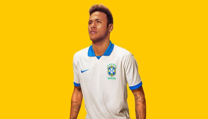 Camisa retrô reserva da Seleção Brasileira 2019-2020 branca com detalhes azuis. A camisa possui gola polo azul, detalhe triangular azul na parte frontal, logo da Nike azul no lado direito do peito e nas bordas das mangas, detalhe verde com linha central azul na nuca e a frase "O TEAM BRASILEIRO" dentro da gola em azul.