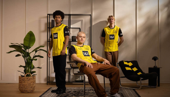 Camisa titular do Borussia Dortmund 2024/25 da Puma. A camisa é amarela com mangas pretas e gola redonda. Quatro listras verticais pretas grossas cruzam o peito, acompanhadas por duas linhas mais finas em seu entorno. O escudo do clube está no lado esquerdo do peito, enquanto o logo da Puma aparece em preto no lado direito e em amarelo nas mangas. O logo da 1&1 aparece dentro de um quadrado preto no espaço master. 