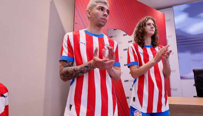 Camisa titular da Seleção Paraguaia Puma 2024-2025 em cores branca, vermelha e azul. A camisa apresenta listras verticais albirrojas, com linhas diagonais azuis nas listras vermelhas. A gola é redonda com a parte frontal azul royal e a nuca branca, enquanto os punhos são azuis.