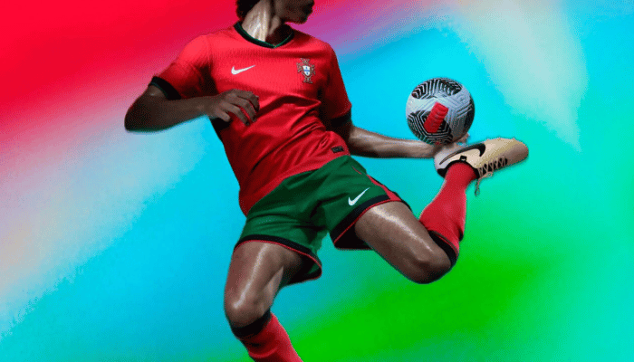 camisa titular da seleção portuguesa 2024-2025. A camisa é predominantemente vermelha com gola preta e contorno verde. Os punhos também são pretos com contorno verde. O escudo da Federação Portuguesa de Futebol está bordado no lado esquerdo do peito e o logotipo da Nike está bordado no lado direito do peito. O modelo está sorrindo e olhando para a câmera.