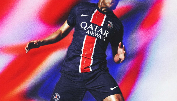 Camisa titular masculina do PSG para a temporada 2024-2025. A camisa é azul marinho com uma faixa vertical vermelha com bordas brancas no estilo "Hechter". O design apresenta um efeito pincelado inspirado na arte urbana da Grande Paris. A gola é redonda e azul marinho, com o nome "PARIS SAINT-GERMAIN" escrito na parte interna. 