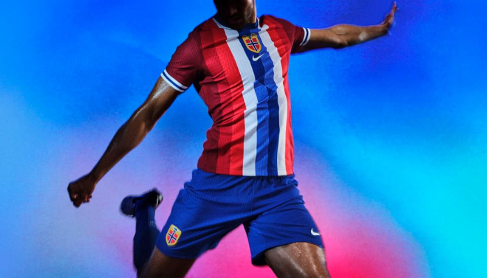 Camisa titular da Seleção Norueguesa Nike 2024/25. A camisa é predominantemente vermelha com listras verticais em três tons de vermelho nas laterais, branco no centro e dois tons de azul entrelaçados. As mangas são grenás e os punhos são brancos com uma faixa azul centralizada. A gola é redonda e azul. No interior da gola, está escrita a frase "Mais fortes juntos".