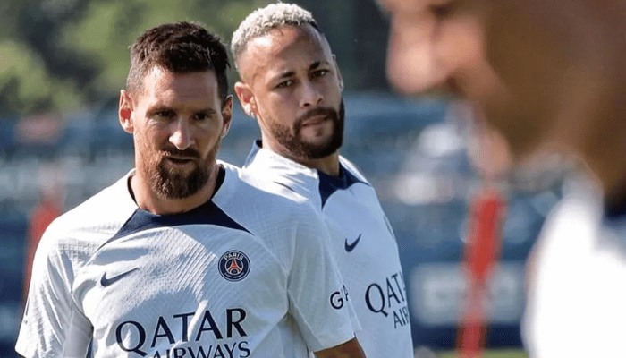 Imagem de uma camisa de treino branca com detalhes em azul marinho e vermelho. A camisa tem o escudo do PSG no lado esquerdo do peito e o logo da Nike no lado direito. Há um fino painel elástico vermelho próximo à bainha.