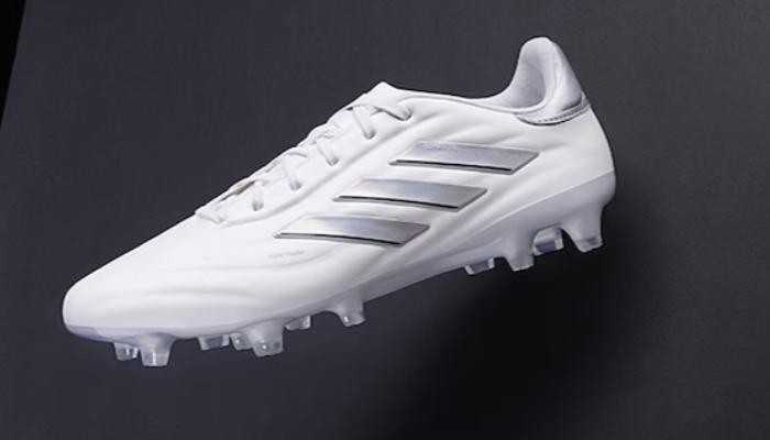 Chuteira Adidas Copa Pure 2+ FG Base White em destaque. A chuteira é predominantemente branca com detalhes em preto e dourado. As Três Listras da Adidas estão presentes na lateral da chuteira em metálico.