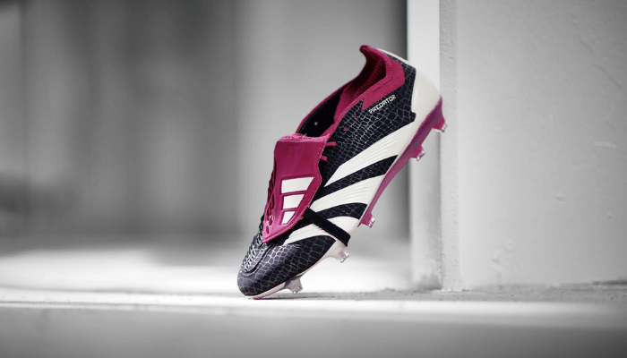 Chuteira Adidas Predator Foldover Tongue FG 30th Anniversary. A chuteira é preta com detalhes em vermelho e branco. A parte superior da chuteira é texturizada e a língua é dobrável. A chuteira está posicionada em um campo de grama.