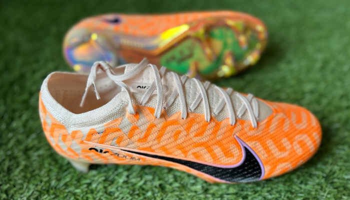  Chuteira Nike Air Zoom Mercurial Vapor 15 Elite FG Pacote United. A chuteira possui um design moderno com a base na cor "guava ice" e um gráfico NU repetido por toda a parte superior. O solado é iridescente e oferece um visual único. A chuteira está apoiada em uma superfície plana.
