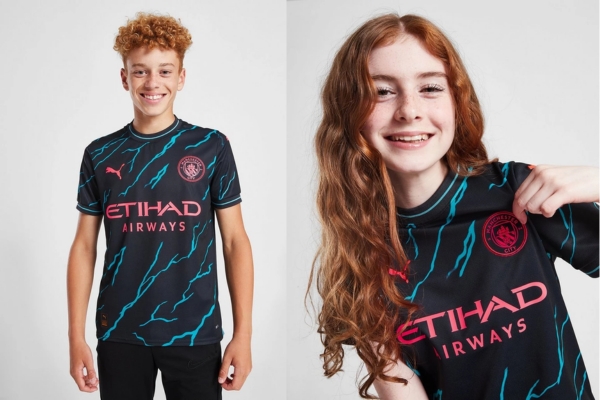 Conjunto infantil Manchester City 2023/24 Puma: azul marinho e vermelho elétrico com padrão de faísca. Detalhes em rosa neon no brasão do clube e logotipos dos patrocinadores