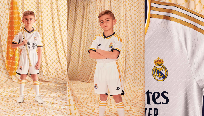 Conjunto infantil titular do Real Madrid Adidas 2023-2024. Camisa branca com gola V em azul marinho, branco e dourado, escudo do Real Madrid no lado esquerdo do peito, logo da Adidas no lado direito e frase "¡Hala Madrid!" na nuca. Calções brancos completam o uniforme.