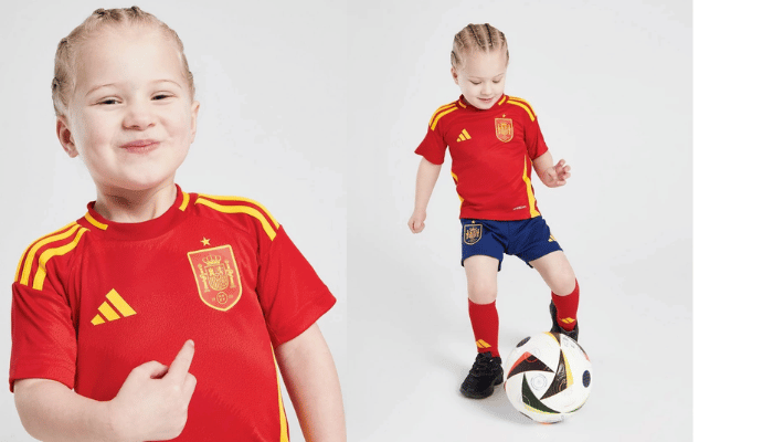 Conjunto infantil da Seleção Espanhola Adidas 2024-2025. Camisa vermelha "Fúria" com detalhes em amarelo e calção azul royal. Menino vestindo o uniforme e vibrando com a seleção.