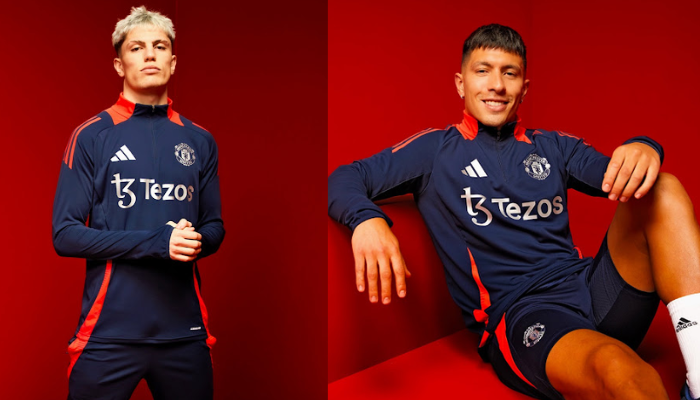 Conjunto de treino oficial do Manchester United Adidas 2024-25 em close-up. Apresenta as cores azul marinho e laranja, detalhes em prata metálica, o brasão do Manchester United e o logotipo da Adidas. O modelo veste uma jaqueta com zíper frontal e punhos com polegar, e uma calça esportiva com o logo do clube.