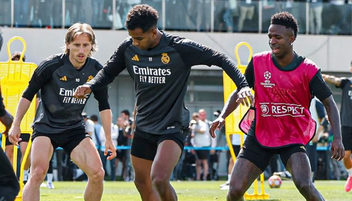 Jaqueta de Treino Real Madrid Adidas UCL 2024 Cinza e Preta. A jaqueta é cinza com detalhes em preto, com o escudo do Real Madrid e o logotipo da Adidas. O modelo está vestindo a jaqueta em um campo de futebol.