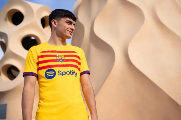 Quarta camisa Barcelona Nike 2023-2024 masculina em amarelo vibrante, listras vermelhas ousadas e toques de azul. Inspirada na braçadeira de capitão de Sergio Busquets, gola redonda amarela, punhos blaugrana e Swoosh da Nike em azul escuro.