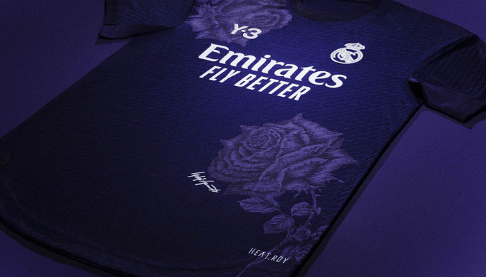 quarta camisa do Real Madrid 2024/25 na cor roxa. A camisa possui duas rosas em tom sobre tom, uma na parte superior direita e outra na parte inferior esquerda com a assinatura de Yohji Yamamoto. A gola é redonda, as mangas e punhos são da mesma cor roxa da camisa. O escudo do Real Madrid é aplicado em branco no lado esquerdo do peito, enquanto o logo da Y-3 e o patrocínio da Emirates também são brancos.