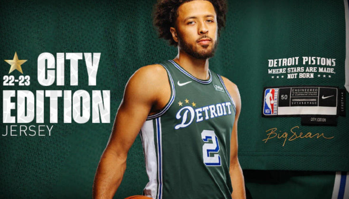 Uma regata de basquete verde, branca e azul com o logotipo do Detroit Pistons no peito. Acima do logotipo, três estrelas representam os três campeonatos da NBA da equipe. A palavra "DETROIT" está escrita em letras brancas na parte superior da frente da regata.