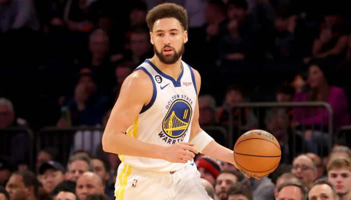 regata branca, azul e amarela com o logotipo do Golden State Warriors. A regata tem o número 30 nas costas e o nome "CURRY" acima do número. O modelo na foto está sorrindo e segurando uma bola de basquete.