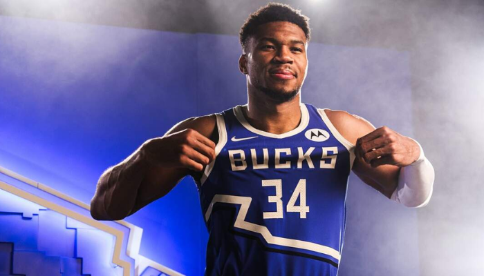 Regata City Edition 2024-25 do Milwaukee Bucks. Em azul e branco, destaca a fronteira de Wisconsin e o nome do time, homenageando Milwaukee e seus fãs, inspirada no histórico apelido Ojibwe "ominowakiing" (local de encontro perto da água).