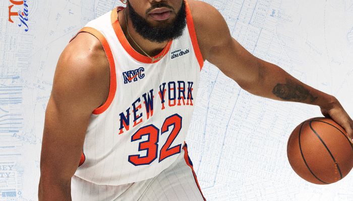 Regata City Edition 2024-25 do New York Knicks. A camisa branca apresenta a icônica marca empilhada "New York" em azul sobre laranja, com detalhes em azul royal nas laterais. O logotipo Nike NYC City Attack aparece no peito.