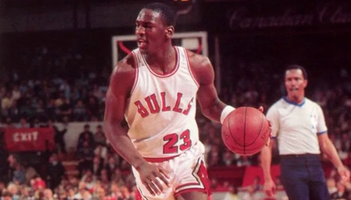 egata titular do Chicago Bulls da temporada 1984-1985, com o icônico número 23 e o logo dos Bulls. A regata é branca com detalhes em vermelho e preto, a mesma usada por Michael Jordan em sua temporada de estreia na NBA