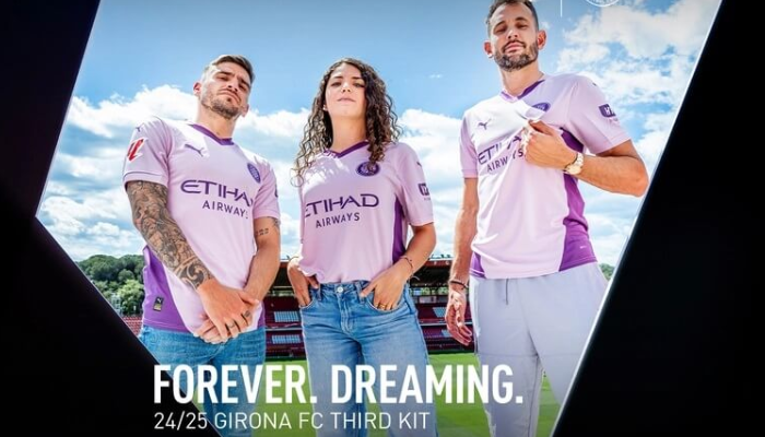 A terceira camisa do Girona FC para a temporada 2024-2025, com design moderno e elegante, apresenta a cor lilás como predominante e detalhes em roxo. A gola V, com o vértice em roxo, e a frase "Orgulho de Girona" na nuca, destacam a identidade do clube. 